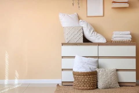Commode with stack of clean bed sheets, baskets and pillows near beige wall i Stock Photos