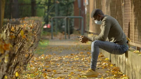 Communication in time of covid - black man types on his mobile sitting at park Stock Footage 143484632
