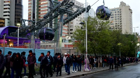 Commuters queue beneath urban gondola line in busy city street scene Stock Footage 327423559
