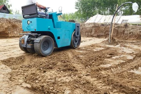 A compact roller is actively compressing dirt on a construction site, where.. Stock Photos