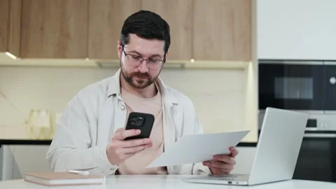 Competent male looking from document to tphone during work process at home Stock-Footage 267184084