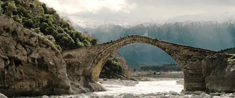 Composition of ancient stone arch bridge over rocky riverbed framed by mountains Stock Footage 328934887