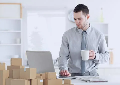 Composition of stack of cardboard boxes and man using laptop at home Stock Photos
