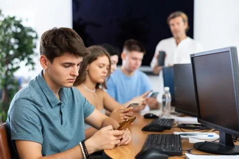 At computer class, young guy student sits with mobile phone and listens Stockfoto's