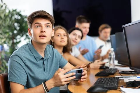 At computer class, young guy student sits with mobile phone and listens Stock Photos