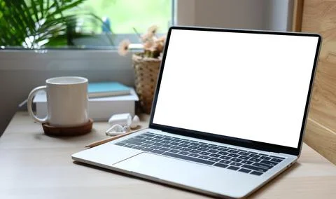 Computer laptop with blank screen and coffee cup on wooden table near window. Stock Photos
