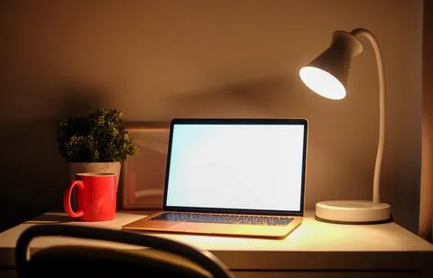 Computer laptop with blank screen, coffee cup and lamp on white table in dark Stock Photos