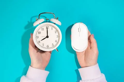 Computer mouse and alarm clock in female hands on a blue background, top view. Stock Photos