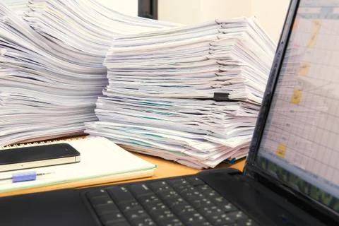 Computer notebook and documents on desk stack up high waiting to be managed Stock Photos