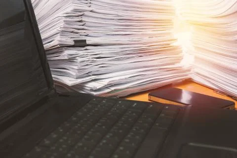 Computer notebook and documents on desk stack up high waiting to be managed Stock Photos