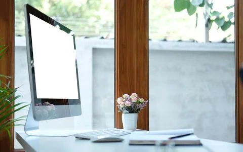 Computer, notebook and flower pot on white table near glass window. Blank scr Stock Photos