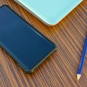 Computer notebook with black smartphone and a blue pencil on a wooden desk. Stock Photos