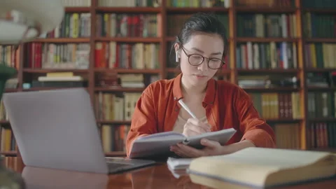 Concentrated asian student writing down notes, taking online lessons in library Video stock 158931606