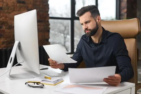Concentrated caucasian guy is doing paperwork. Man looking through paper sitting Stock Photos