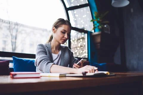 Concentrated female freelancer checking notes and browsing smartphone in cafe Stock Photos