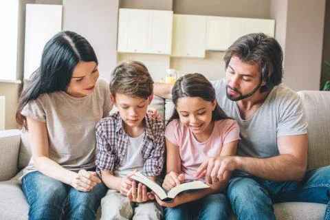 Concentrated kids are sitting on sofa with their parents and reading a book Stock Photos
