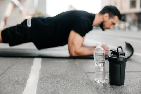Concentrated man doing plank position while exercising on black yoga mat. Water 스톡 사진