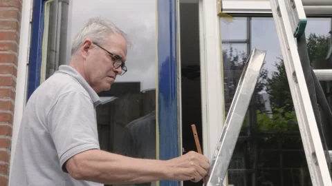 Concentrated man paints window frames in blue color Stock Footage 279991759