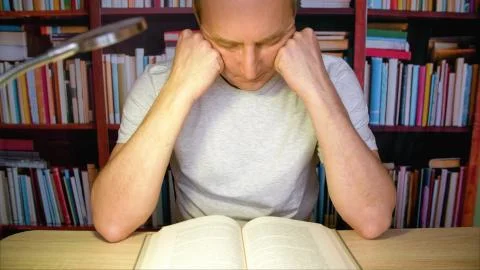 Concentrated man reading book at table with lamp in cozy library at evening Stock Photos