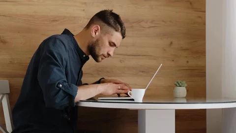 Concentrated man types on laptop while sitting at kitchen table with cup of Stock Footage 342544645