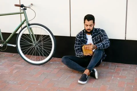 Concentrated man using a tablet while sitting near the bike, outdoors. Stock Photos