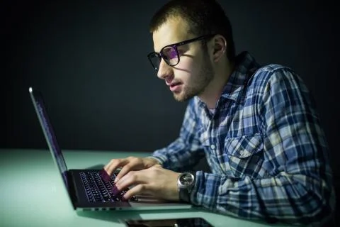 Concentrated young hacker using computer and stealing information in dark roo Stock Photos