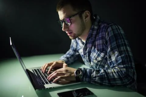 Concentrated young hacker using computer and stealing information in dark roo Foto stock