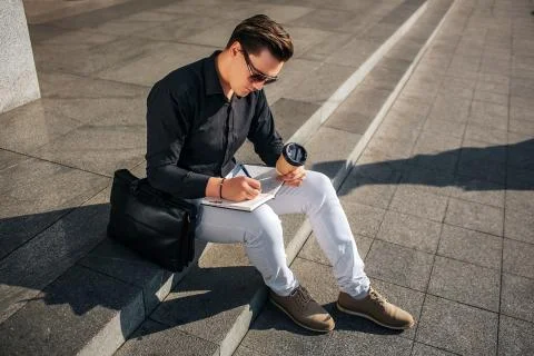 Concentrated young man sit on steps on street and write in notebook. He holds Stock Photos