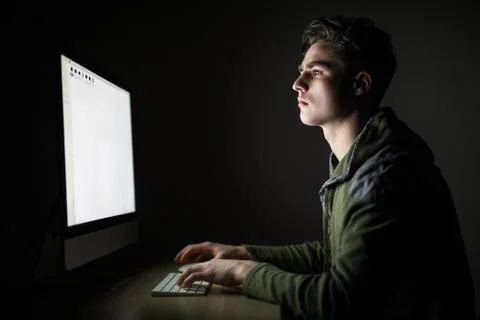 Concentrated young man sitting at the table and using computer in dark room 스톡 사진