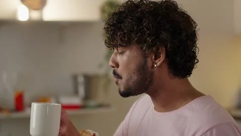 Concept of cheat meal. Young indian man eating big sandwich and drinking Stock Footage 254157084