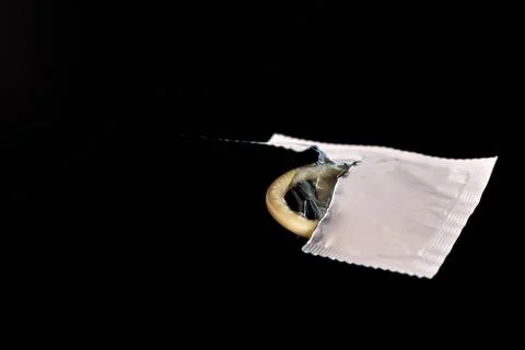 A condom is half pulled out of an open package on a dark background. Side vie Stock Photos