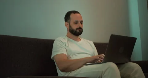Confident brunette guy with a beard in a white T-shirt sits on a gray sofa and Video stock 280653075