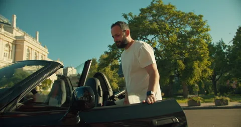 A confident brunette guy in a white T-shirt approaches a black convertible and Stock Footage 280577625