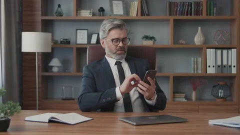 Confident businessman with glasses using his smartphone sitting at table in Vídeos de archivo 220189061
