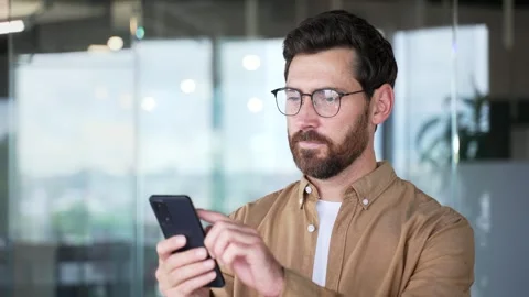 A confident businessman is using browsing mobile phone while standing in a moder Stock Footage 325513053