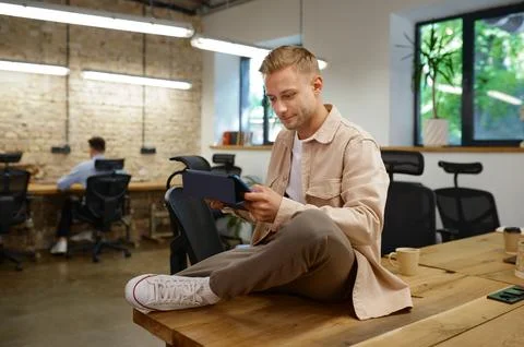 Confident concentrated man creative worker sitting on office table with tablet Stock Photos