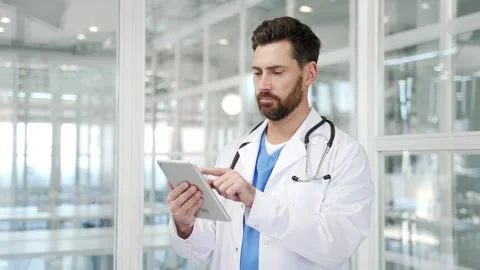 Confident doctor in white coat using digital tablet while standing at workplace  Stock Footage 305693918