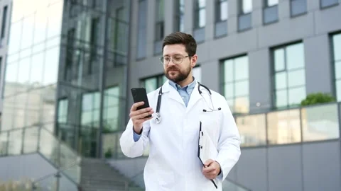 Confident doctor in white coat using mobile phone while walking outside near  库存影片 308098901