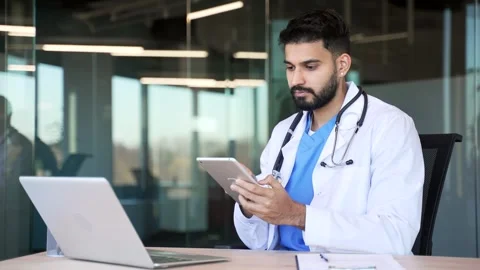 Confident doctor in white coat using digital tablet while sitting at workplace  Stock Footage 320619282