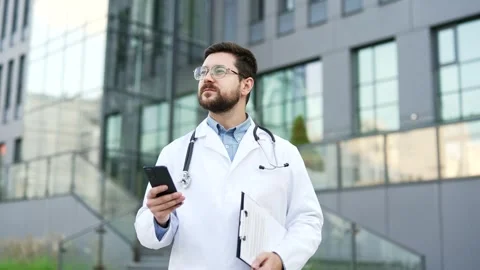 Confident doctor in white coat using mobile phone while standing outside near  Stock Footage 321291496