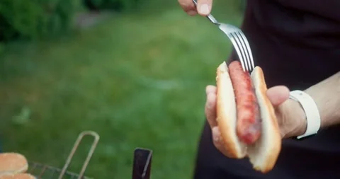 Confident guy in a black apron puts a sausage inside a bun while preparing Stock Footage 283832337