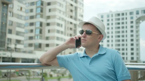 Confident man in hat talking on mobile phone while standing near fence railing Stock Footage 153630183