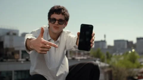 Confident man presenting phone toward camera on rooftop with pointing gesture Stock Footage 345241047