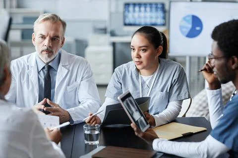 Confident mature doctor and his young female assistant looking at colleague Stock Photos
