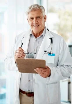 Confident mature male doctor reading patient records on clipboard in hospital or Stock Photos