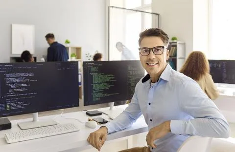 Confident programmer posing by code monitors at desk in bright modern office Stock-Fotos