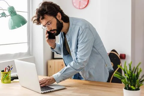 Confident young bearded creative man talking on phone while using laptop Stock Photos