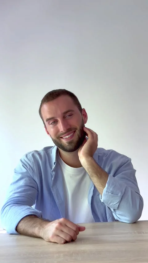 Confident young man with a beard sitting at a wooden desk, smiling and looking Stock Footage 346605177
