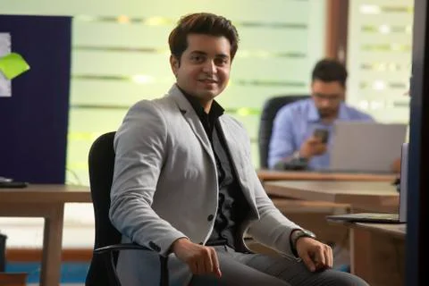 A CONFIDENT YOUNG MAN LOOKING AT CAMERA AND SMILING WITH COLLEAGUE SITTING BEHIN Stock Photos