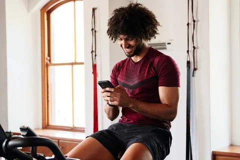 Confident young middle eastern man using mobile phone on exercise bike at the Stock Photos